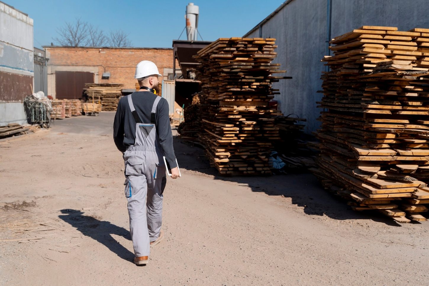 Construction Materials Nicholasville KY 1 - Stratton Lumber - Stratton Lumber Construction worker in a white hard hat and gray overalls walking through a lumberyard with stacked planks nearby