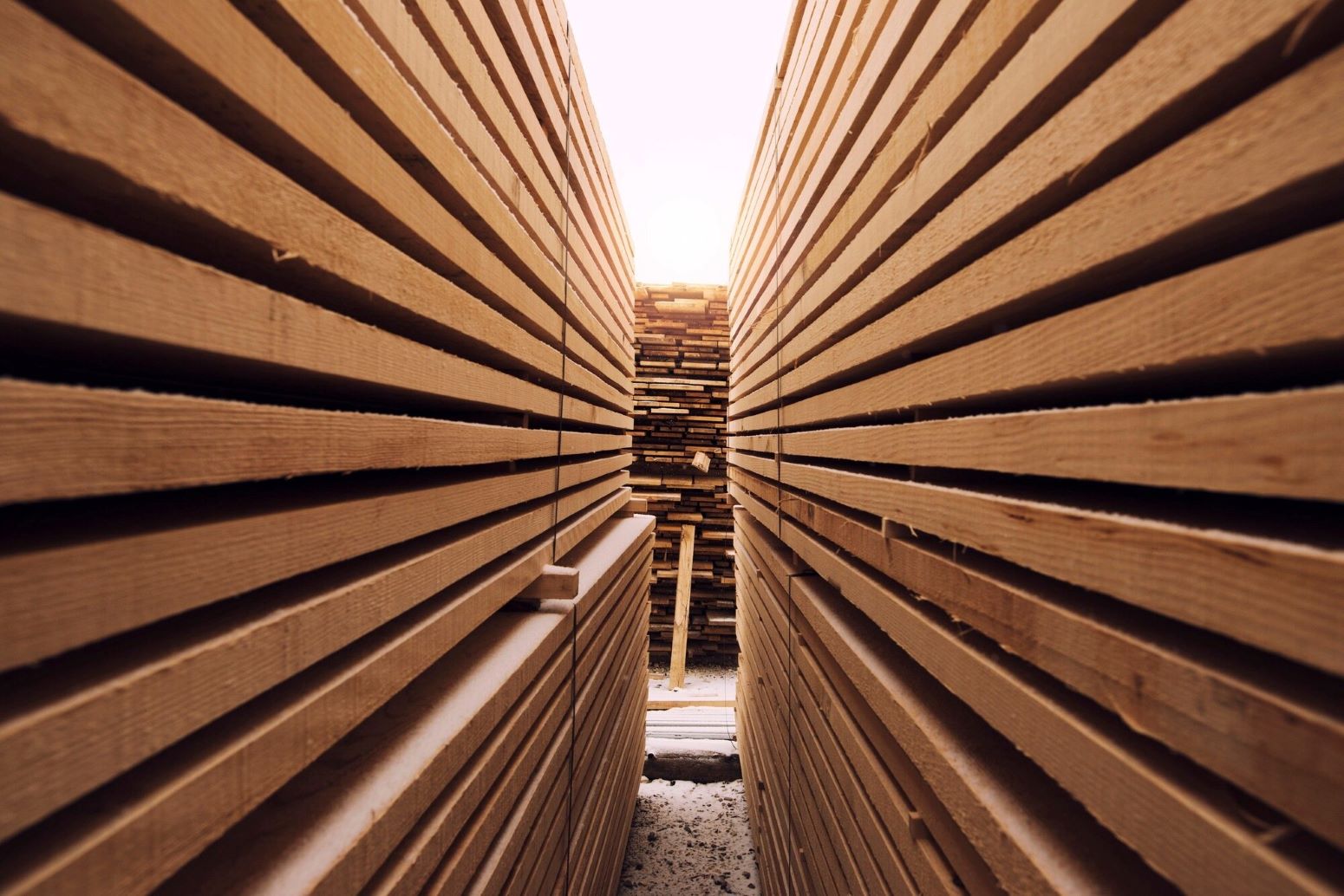 Narrow corridor between tall stacks of wooden planks, sunlight at the far end.
